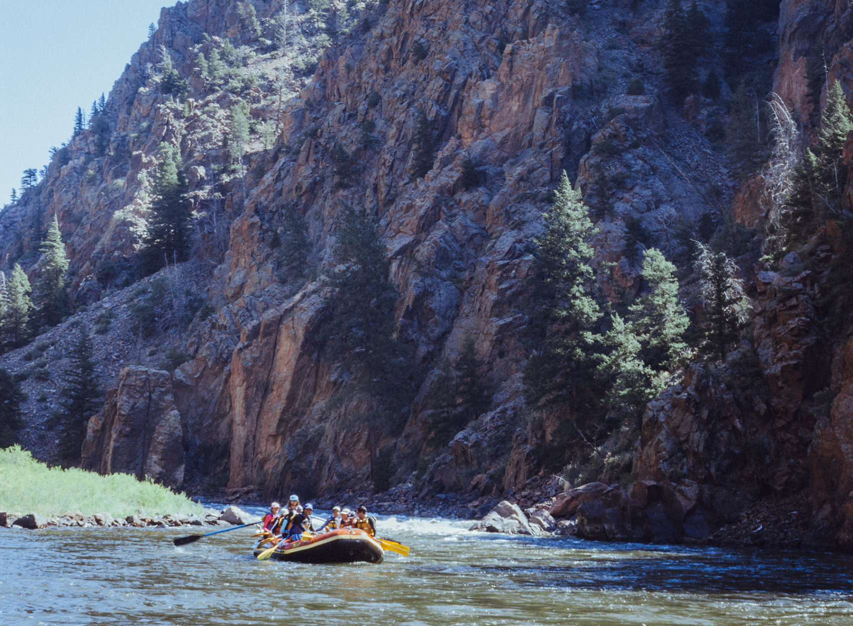 A raft coming down the Colorado River with a steep mountain wall behind them